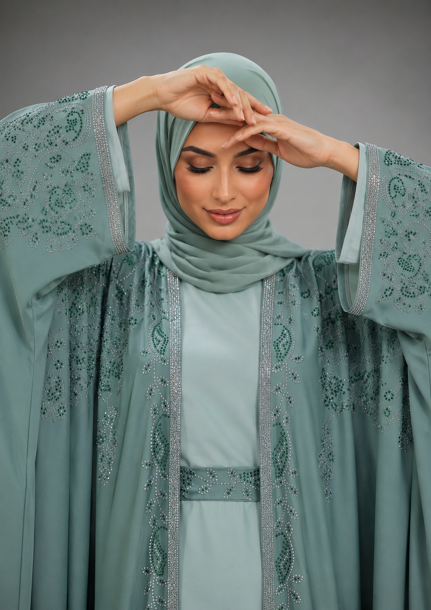 A woman modeling a sage green modest outfit featuring a matching hijab, a light sage green dress, and a sheer embroidered abaya with silver beaded borders, posed against a solid gray studio background.