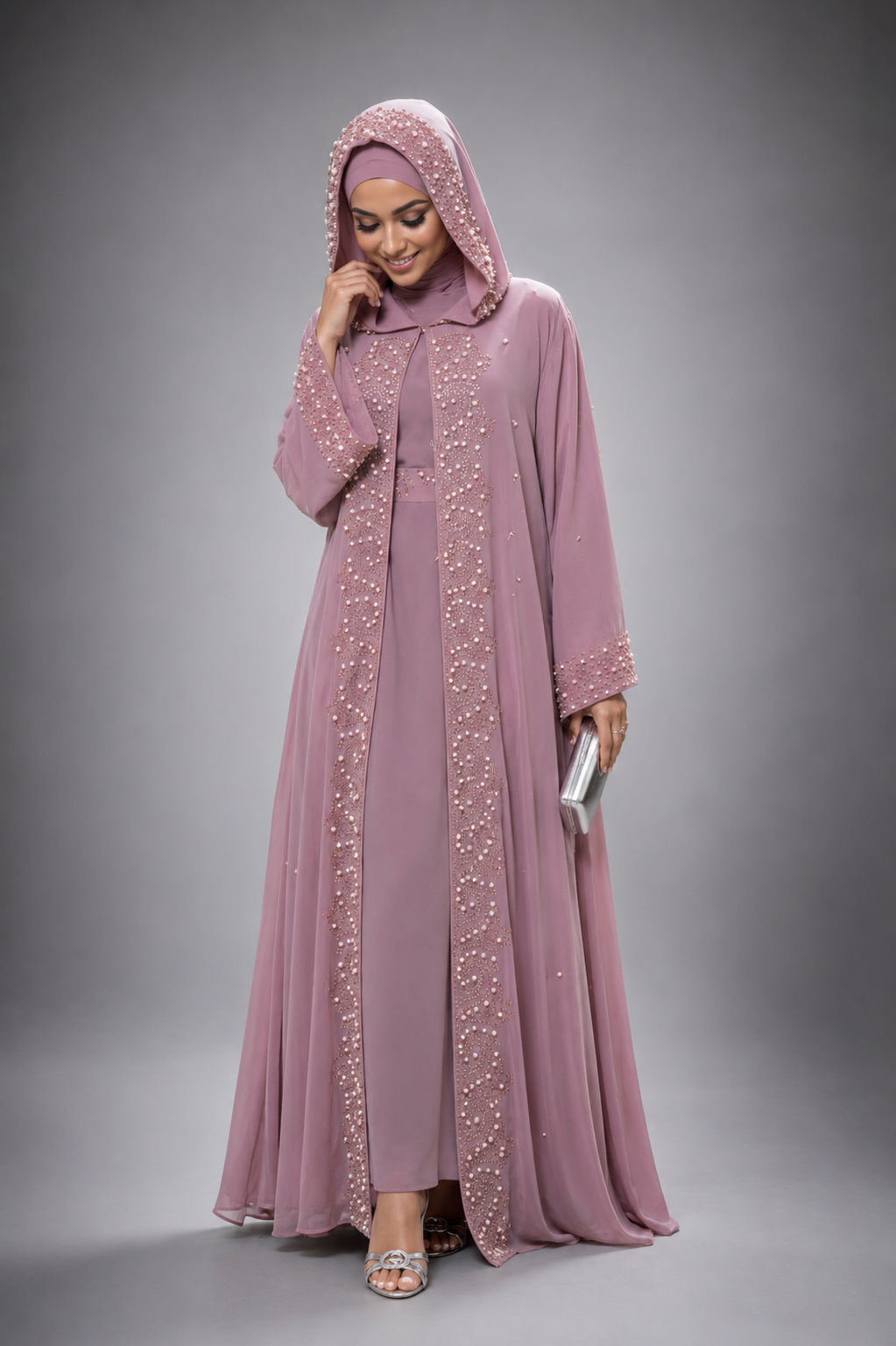 A woman modeling a full-length dusty pink abaya with matching hijab, decorated with sparkling silver beadwork along the front opening and sleeve cuffs, set against a neutral gray studio background.