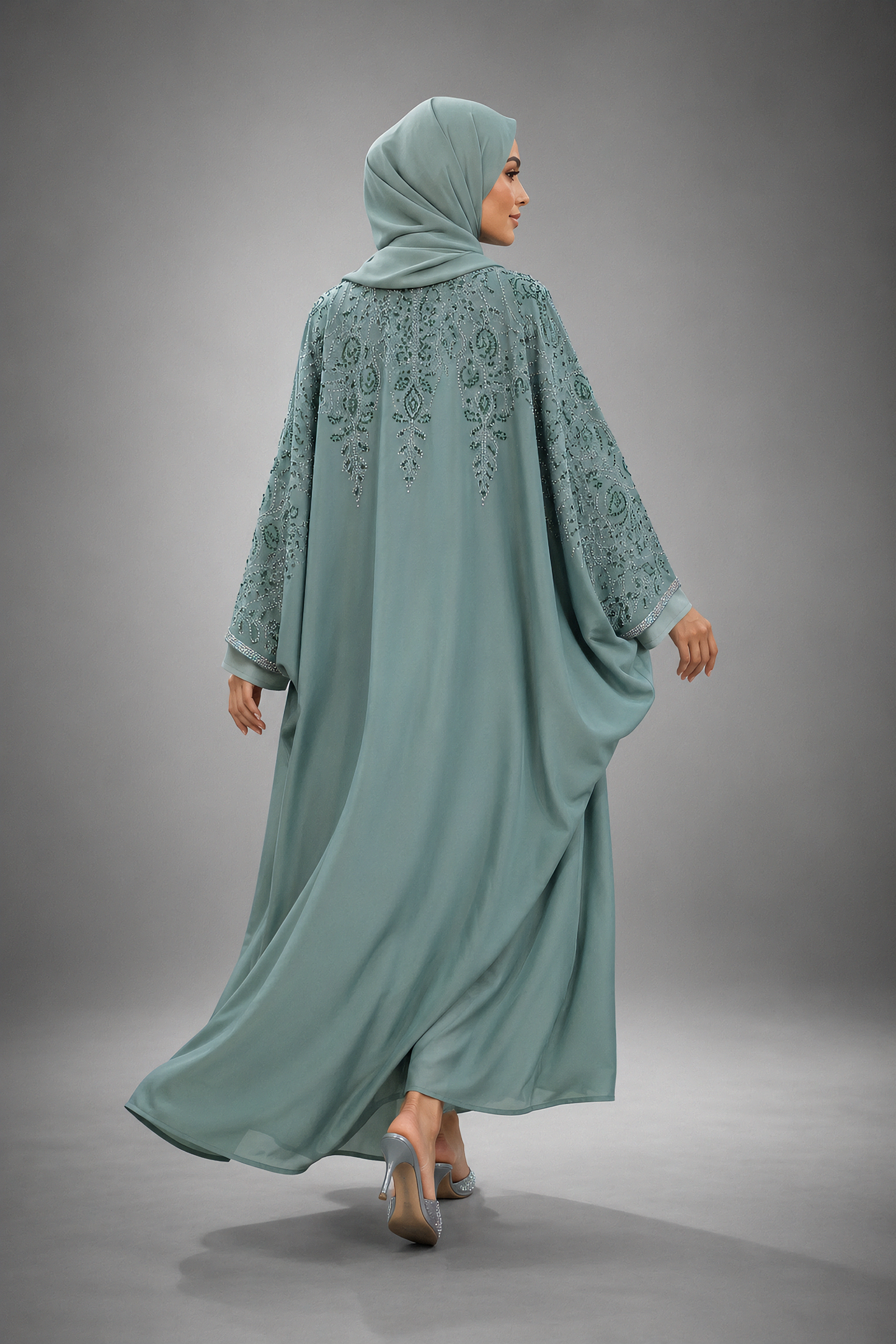 A woman wearing a flowing sage green abaya with floral embroidery on the back and sleeves, paired with a matching hijab, photographed from behind against a neutral gray studio background.
