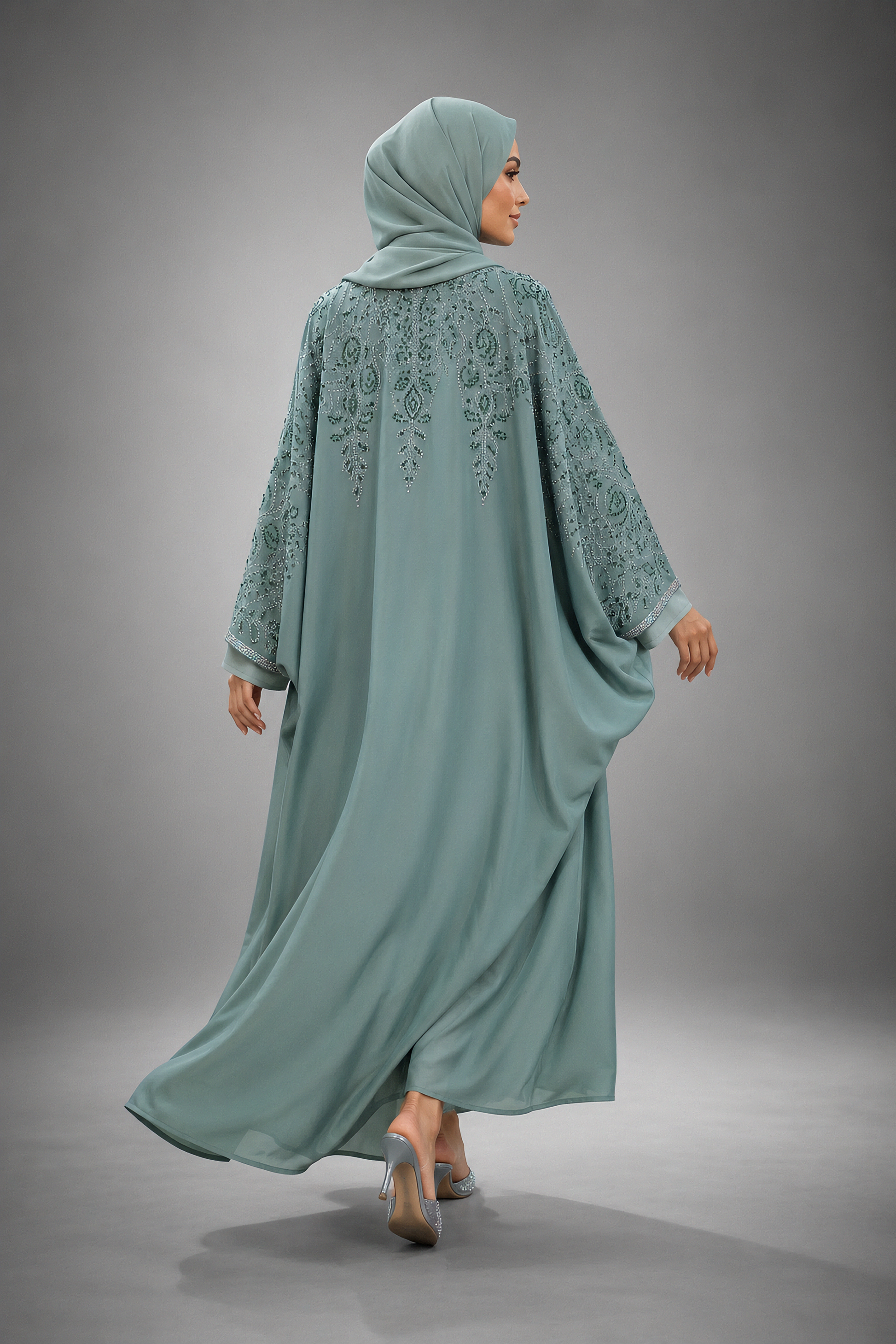 A woman wearing a flowing sage green abaya with floral embroidery on the back and sleeves, paired with a matching hijab, photographed from behind against a neutral gray studio background.