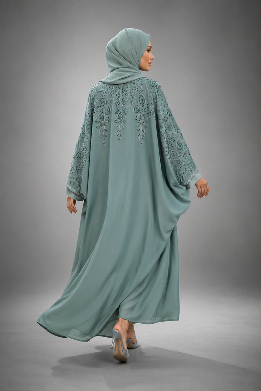 A woman wearing a flowing sage green abaya with floral embroidery on the back and sleeves, paired with a matching hijab, photographed from behind against a neutral gray studio background.