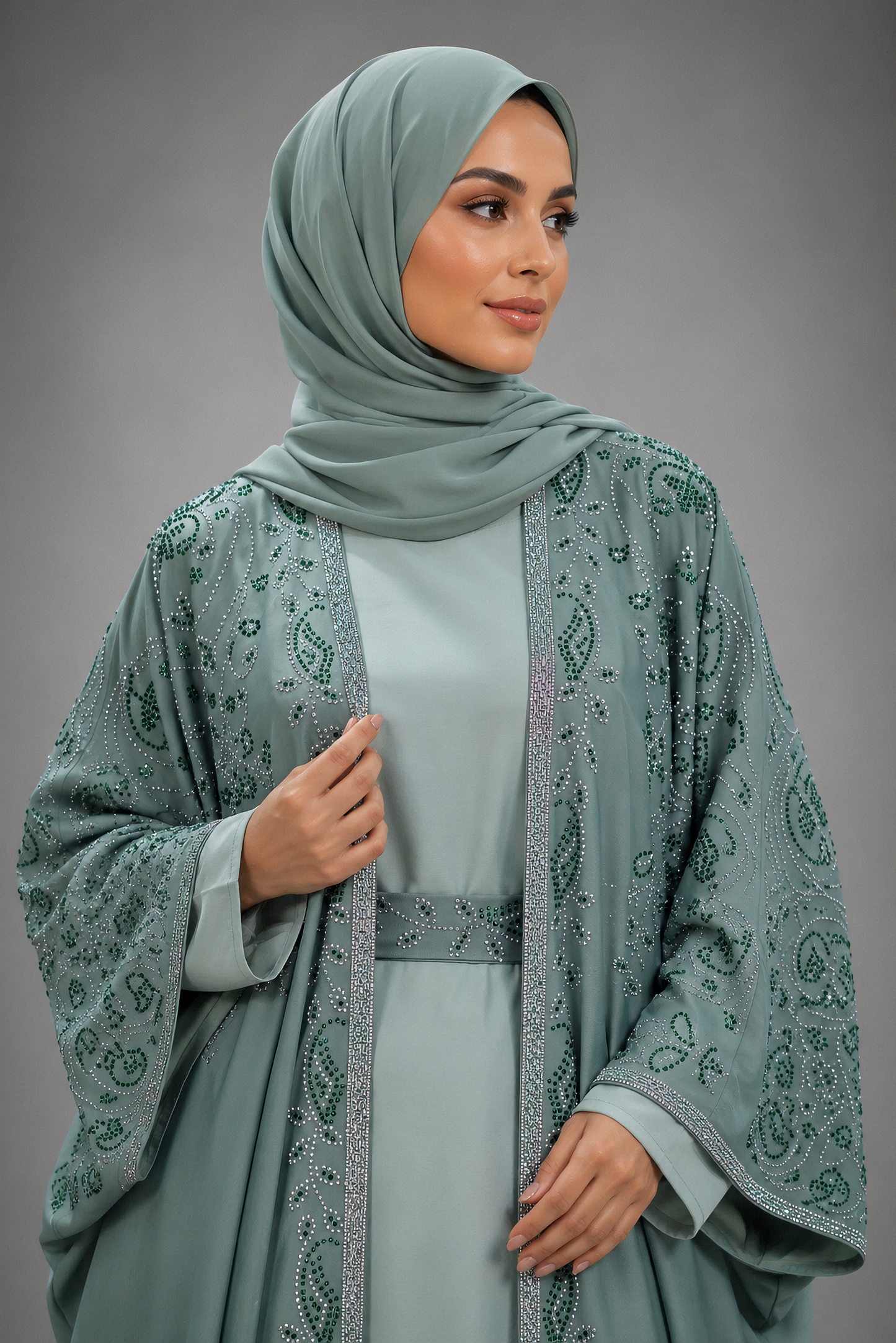 A woman wearing a sage green hijab, matching long-sleeve abaya, and a sheer embroidered outer layer with silver-toned beadwork, posed against a neutral gray studio background.