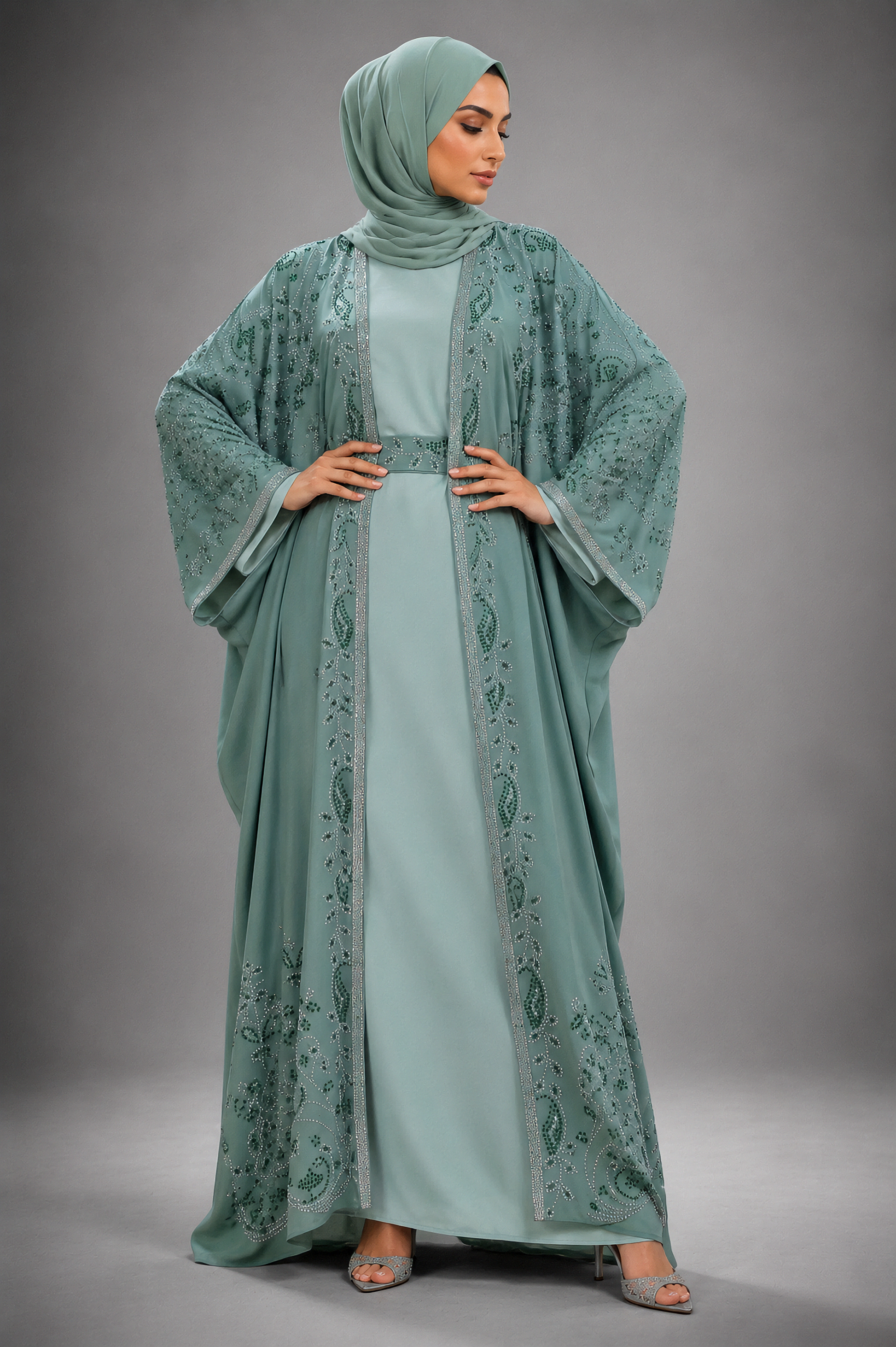 A woman modeling a modest outfit consisting of a sage green abaya with matching embroidered border, a solid sage green inner dress, and a matching sage green hijab, set against a neutral gray studio background.