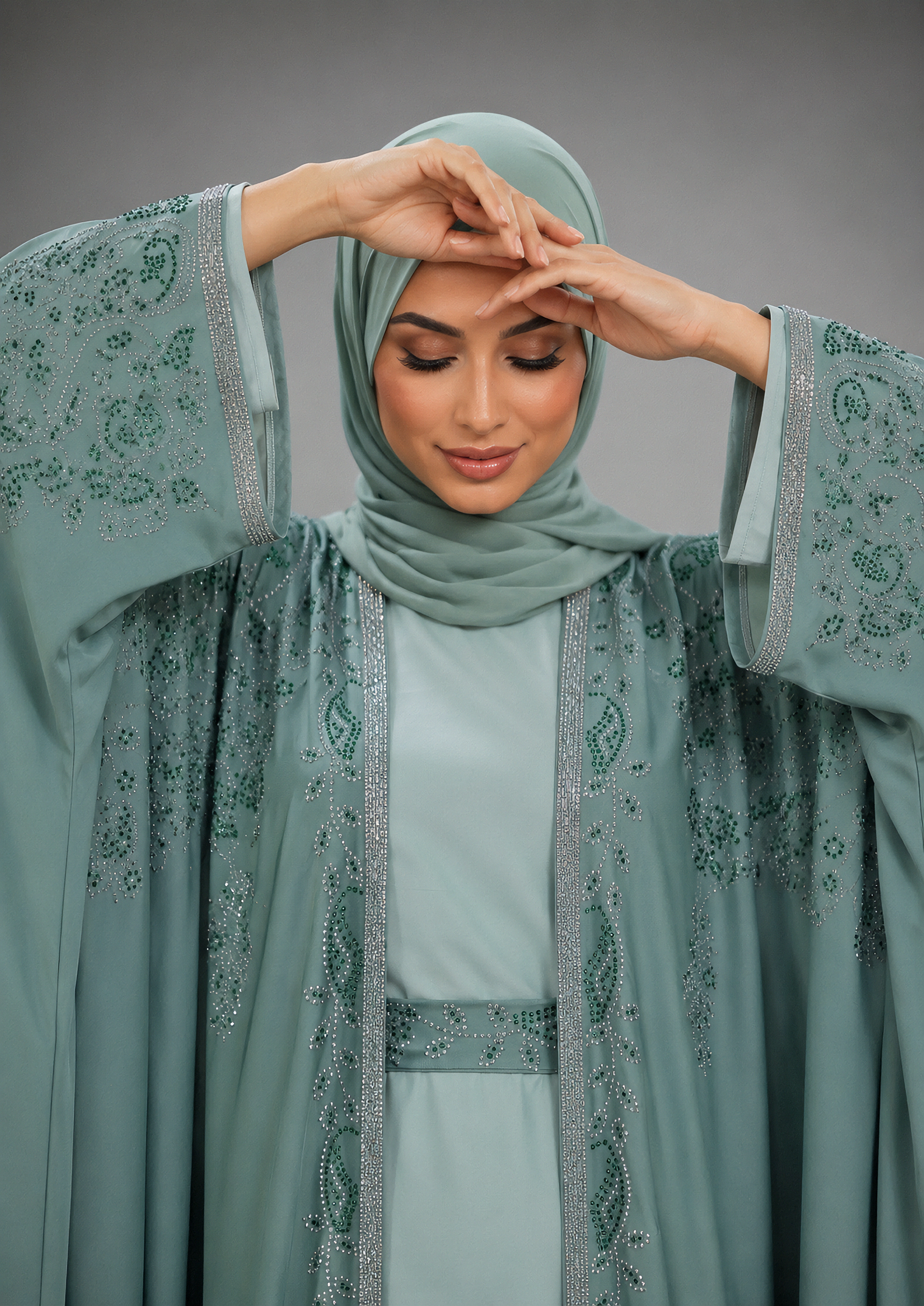 A woman modeling a sage green modest outfit featuring a matching hijab, a light sage green dress, and a sheer embroidered abaya with silver beaded borders, posed against a solid gray studio background.